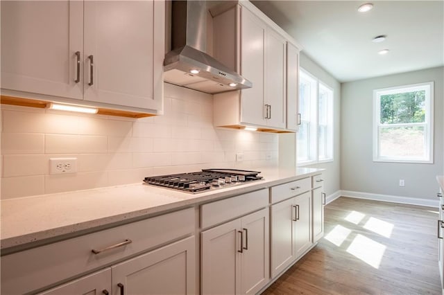 kitchen with light stone counters, wall chimney exhaust hood, stainless steel gas cooktop, light hardwood / wood-style flooring, and white cabinets