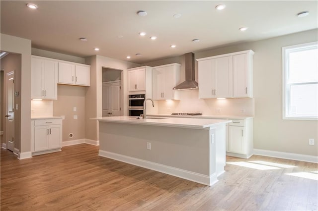 kitchen with light wood-type flooring, an island with sink, white cabinetry, and wall chimney range hood