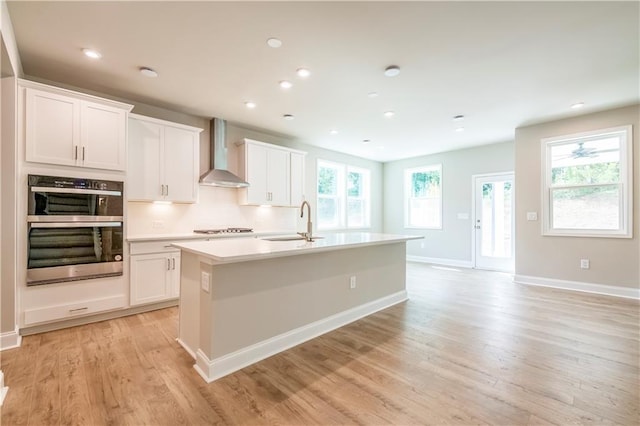 kitchen with a kitchen island with sink, sink, wall chimney exhaust hood, stainless steel double oven, and white cabinetry