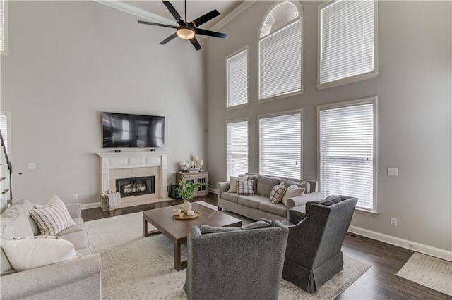 living area featuring baseboards, dark wood finished floors, a fireplace, a towering ceiling, and a ceiling fan