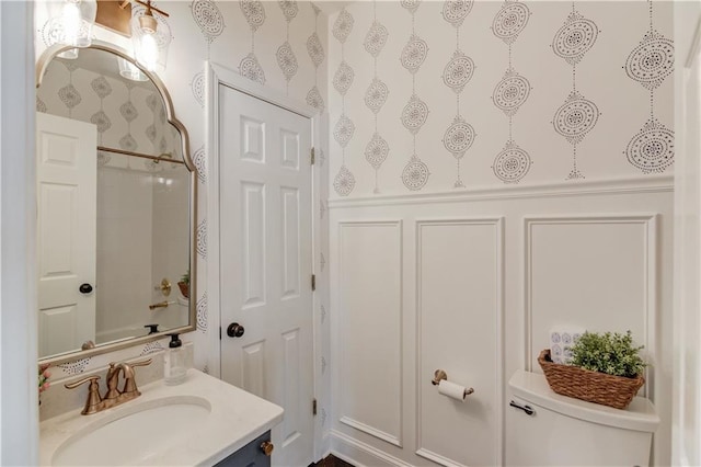 bathroom featuring wallpapered walls, a wainscoted wall, toilet, vanity, and a shower