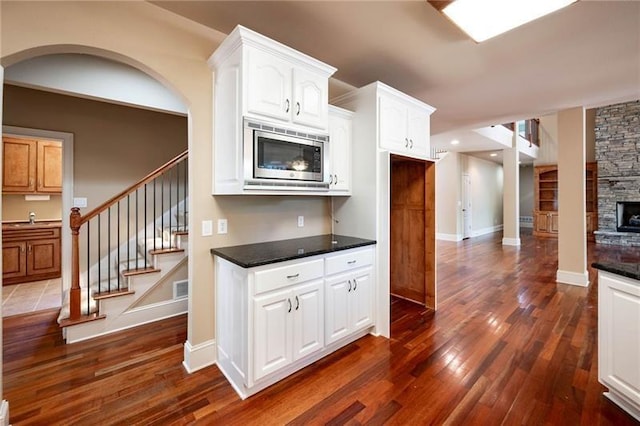 kitchen with a stone fireplace, dark hardwood / wood-style floors, stainless steel microwave, white cabinetry, and dark stone countertops