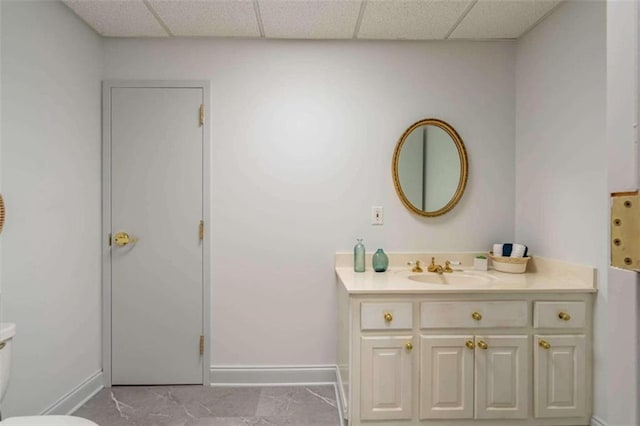bathroom featuring baseboards, a drop ceiling, vanity, and toilet