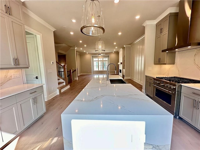 kitchen featuring high end stainless steel range oven, light wood-type flooring, ornamental molding, wall chimney range hood, and an island with sink