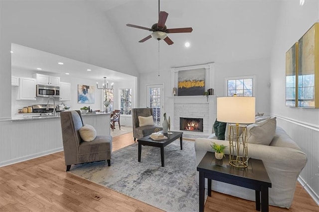 living room featuring a brick fireplace, light wood-type flooring, ceiling fan with notable chandelier, and high vaulted ceiling