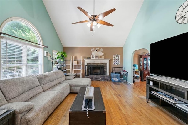 living room with ceiling fan, a stone fireplace, high vaulted ceiling, and light wood-type flooring