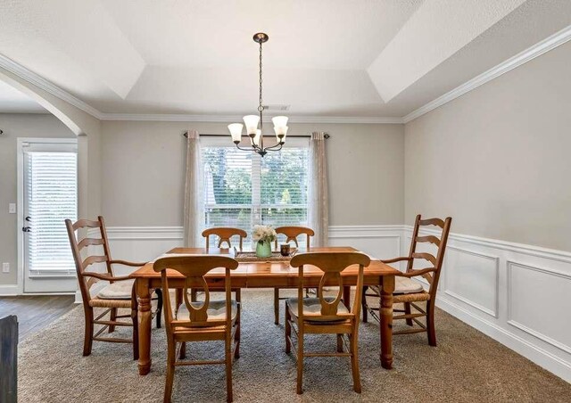 carpeted dining space featuring a chandelier, a wainscoted wall, arched walkways, ornamental molding, and a raised ceiling