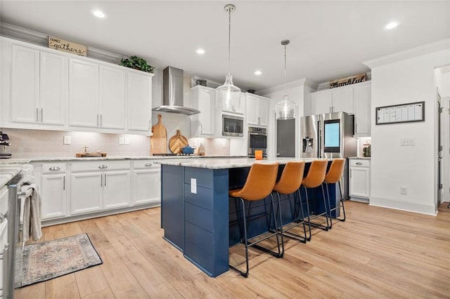 kitchen featuring a center island, wall chimney exhaust hood, stainless steel appliances, pendant lighting, and light hardwood / wood-style floors