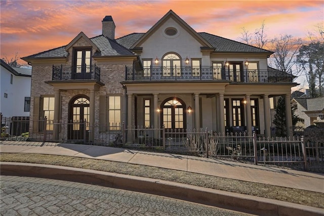 french provincial home featuring a fenced front yard, french doors, brick siding, a chimney, and a balcony