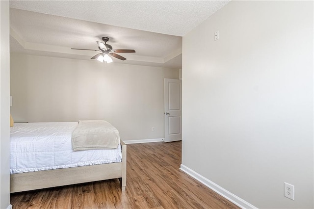bedroom featuring baseboards, wood finished floors, a tray ceiling, a textured ceiling, and a ceiling fan