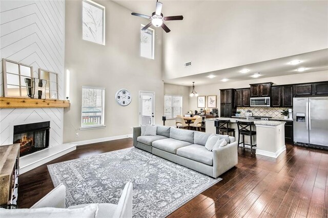 living room with ceiling fan, dark wood-type flooring, a towering ceiling, and a fireplace