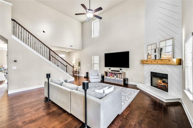 living room with a towering ceiling, a fireplace, ceiling fan, and dark wood-type flooring