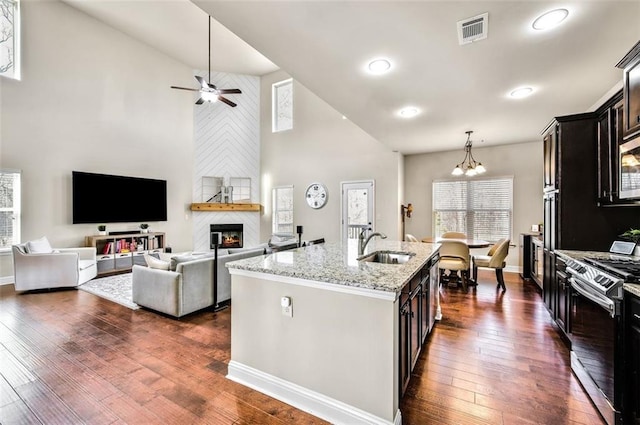 kitchen featuring a large fireplace, a kitchen island with sink, stainless steel range, light stone countertops, and dark hardwood / wood-style floors