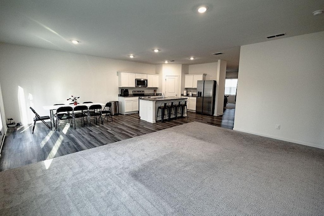 kitchen featuring stainless steel appliances, dark colored carpet, dark countertops, visible vents, and an island with sink