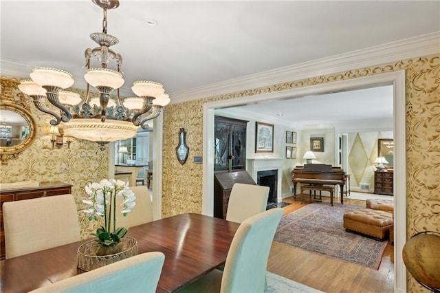 dining area featuring a fireplace with flush hearth, a chandelier, wallpapered walls, ornamental molding, and wood finished floors