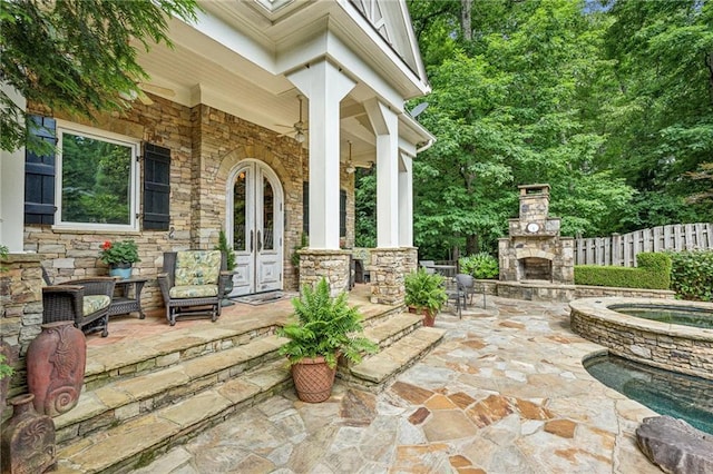 view of patio with an in ground hot tub, an outdoor stone fireplace, fence, a ceiling fan, and french doors