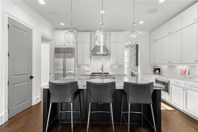 kitchen featuring white cabinets, an island with sink, wall chimney exhaust hood, dark wood-style flooring, and built in fridge