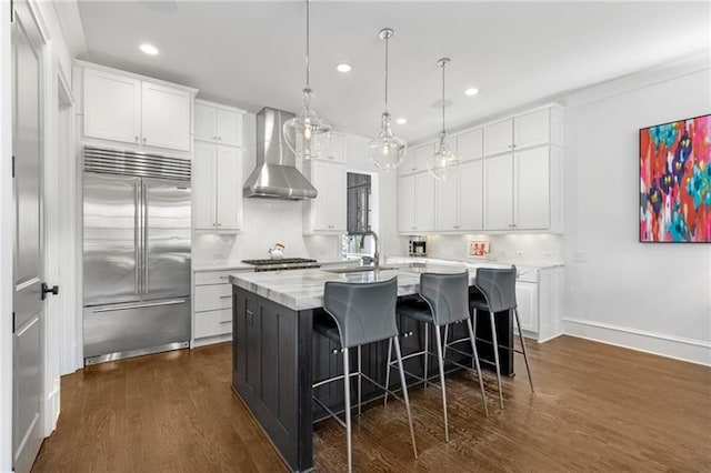 kitchen featuring white cabinets, dark wood finished floors, an island with sink, wall chimney exhaust hood, and built in refrigerator