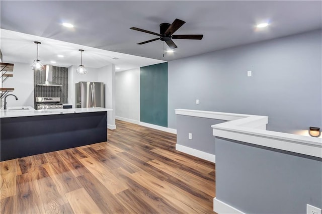 kitchen with light countertops, wall chimney exhaust hood, stainless steel appliances, a ceiling fan, and dark wood finished floors
