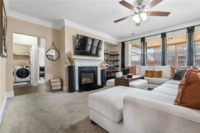 carpeted living area featuring a fireplace with flush hearth, ornamental molding, washing machine and dryer, baseboards, and ceiling fan