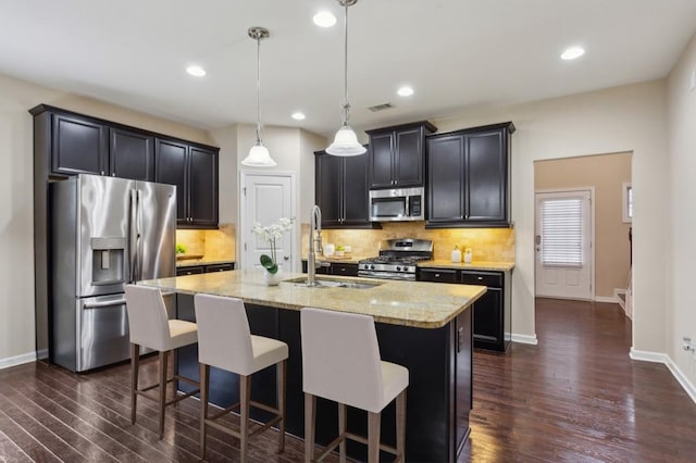 kitchen with light stone countertops, dark wood-type flooring, stainless steel appliances, decorative light fixtures, and a kitchen island with sink