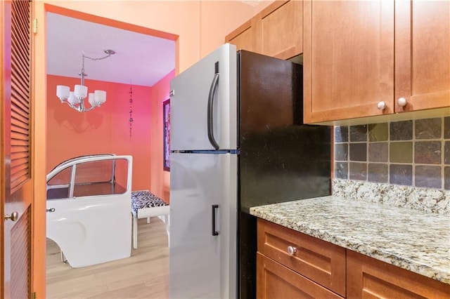kitchen featuring light stone countertops, a notable chandelier, light wood-style floors, freestanding refrigerator, and backsplash