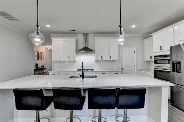 kitchen featuring a center island with sink, wall chimney range hood, white cabinets, and stainless steel appliances