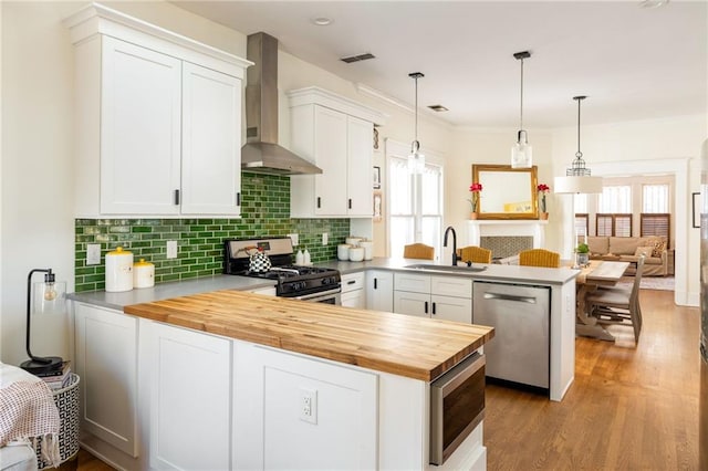 kitchen with wall chimney exhaust hood, a peninsula, stainless steel appliances, wooden counters, and a sink