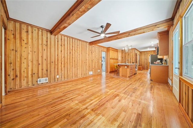 unfurnished living room featuring ceiling fan with notable chandelier, beam ceiling, light wood-type flooring, and wooden walls