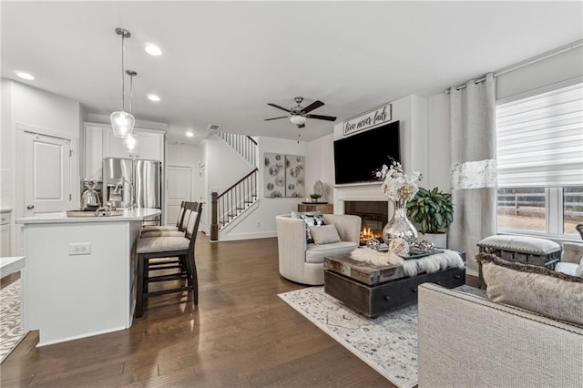 living room with ceiling fan, recessed lighting, dark wood-style flooring, a lit fireplace, and stairway
