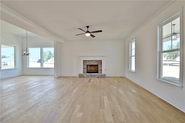 unfurnished living room with ceiling fan with notable chandelier, a stone fireplace, light wood-type flooring, and crown molding