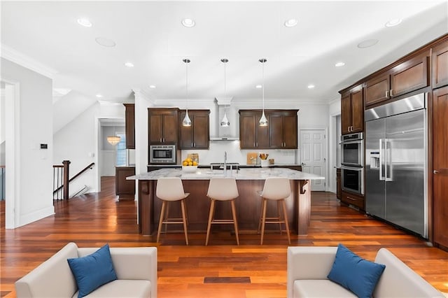 kitchen featuring wall chimney exhaust hood, stainless steel appliances, light stone counters, pendant lighting, and a kitchen island with sink