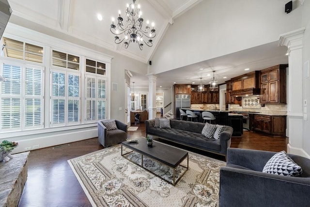 living room with ornate columns, dark wood-type flooring, an inviting chandelier, high vaulted ceiling, and crown molding