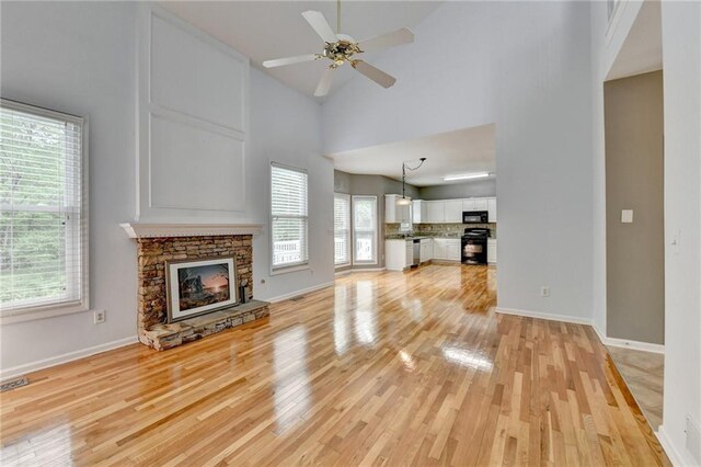 unfurnished living room featuring light wood finished floors, baseboards, ceiling fan, a stone fireplace, and high vaulted ceiling