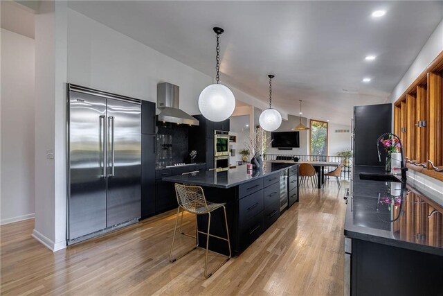 kitchen featuring a kitchen breakfast bar, stainless steel appliances, wall chimney range hood, decorative light fixtures, and light hardwood / wood-style flooring