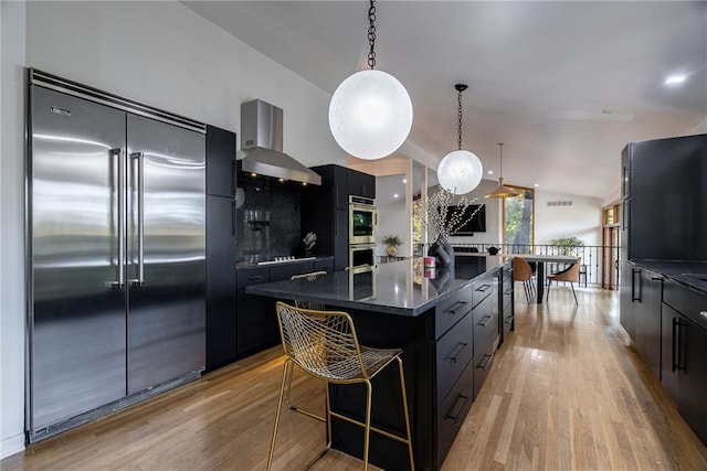 kitchen featuring a spacious island, wall chimney range hood, hanging light fixtures, vaulted ceiling, and appliances with stainless steel finishes