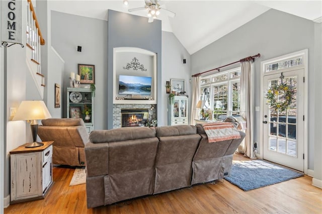 living area featuring high vaulted ceiling, a brick fireplace, stairway, a ceiling fan, and light wood-style floors