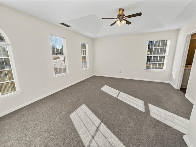 empty room featuring a tray ceiling, dark colored carpet, visible vents, a ceiling fan, and baseboards