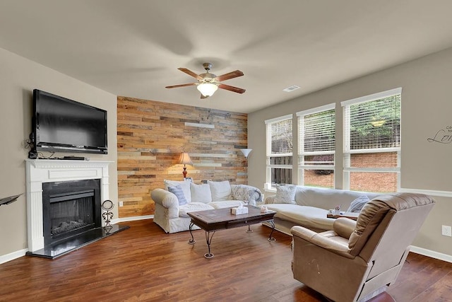living room with dark wood-style flooring, a fireplace with raised hearth, wooden walls, a ceiling fan, and visible vents