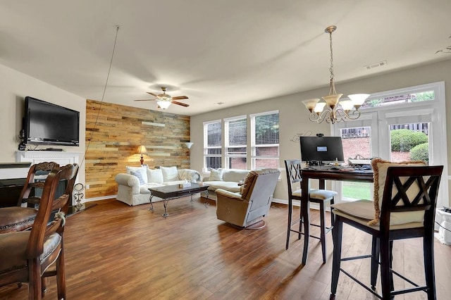 living area featuring visible vents, baseboards, wooden walls, dark wood-type flooring, and ceiling fan with notable chandelier