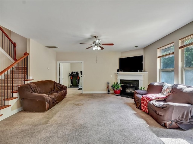 carpeted living room featuring a fireplace with flush hearth, visible vents, stairs, and ceiling fan