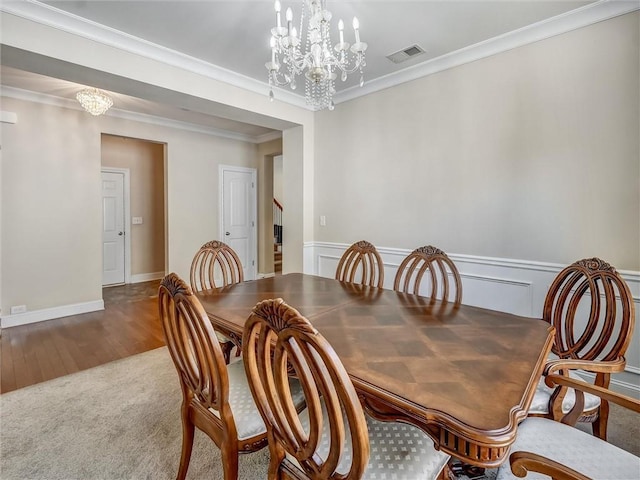 carpeted dining area with wood finished floors, an inviting chandelier, a wainscoted wall, ornamental molding, and visible vents