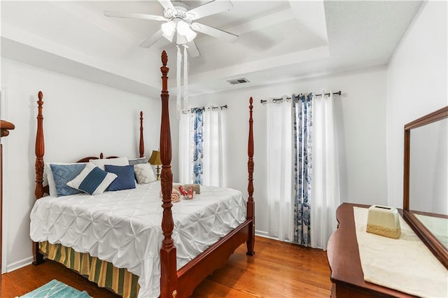 bedroom with a ceiling fan, wood finished floors, visible vents, baseboards, and a tray ceiling