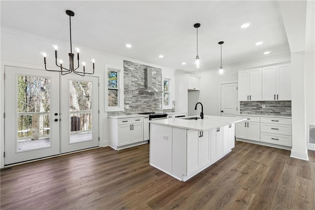 kitchen with wall chimney range hood, white cabinetry, sink, and a center island with sink