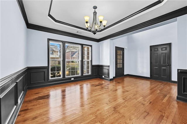 empty room featuring a raised ceiling, wainscoting, ornamental molding, an inviting chandelier, and light wood-type flooring