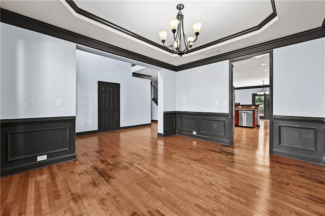 empty room with ornamental molding, wood finished floors, an inviting chandelier, a tray ceiling, and a decorative wall