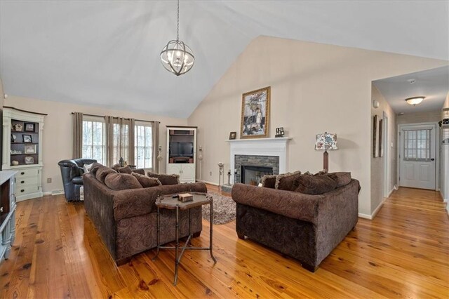 living area featuring baseboards, high vaulted ceiling, a stone fireplace, light wood-style floors, and a notable chandelier