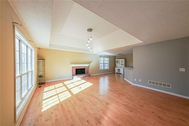 unfurnished living room featuring a healthy amount of sunlight, a tray ceiling, a brick fireplace, and light hardwood / wood-style floors