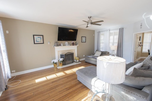 living room with baseboards, light wood-style floors, a fireplace with raised hearth, and a ceiling fan