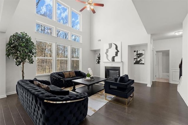 living room featuring ceiling fan, a towering ceiling, wood finished floors, baseboards, and a glass covered fireplace